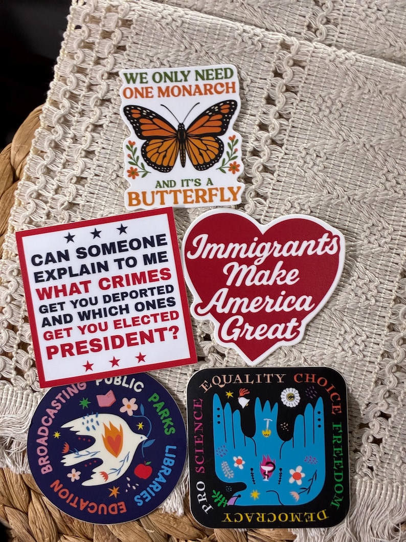 Op de afbeelding: Een verzameling kleurrijke stickers met diverse ontwerpen en tekst. E&eacute;n sticker toont een monarchvlinder, een andere een hart met de tekst "Immigrants Make America Great". Andere stickers bevatten teksten over politiek en sociale kwesties.