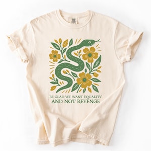 May include: Cream-colored t-shirt featuring a green snake design intertwined with yellow flowers and green leaves. The text "BE GLAD WE WANT EQUALITY AND NOT REVENGE" is printed below the graphic. The shirt has rolled-up sleeves.
