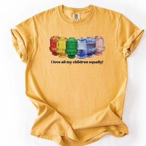 May include: Mustard yellow t-shirt featuring a row of six colorful glass jars. The jars are red, yellow, green, blue, and pink. The text "I love all my children equally!" is printed below the jars.