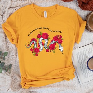 May include: Mustard yellow t-shirt featuring a white snake design intertwined with red roses. The shirt has the text "Be glad we only want equality, and not revenge" in a curved font above the snake design. The shirt is made of a soft material.