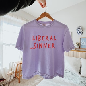 May include: A light purple t-shirt with the words "LIBERAL SINNER" in red lettering. The shirt is hanging on a wooden hanger. The background includes a bed, a small table, and a window.