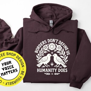 Pode incluir: Um moletom vinho com um gr&aacute;fico branco que diz "Borders Don't Define Us, Humanity Does". O design inclui dois p&aacute;ssaros, flores e folhas. &Agrave; esquerda, h&aacute; um adesivo amarelo com o texto "Your Voice Matters".