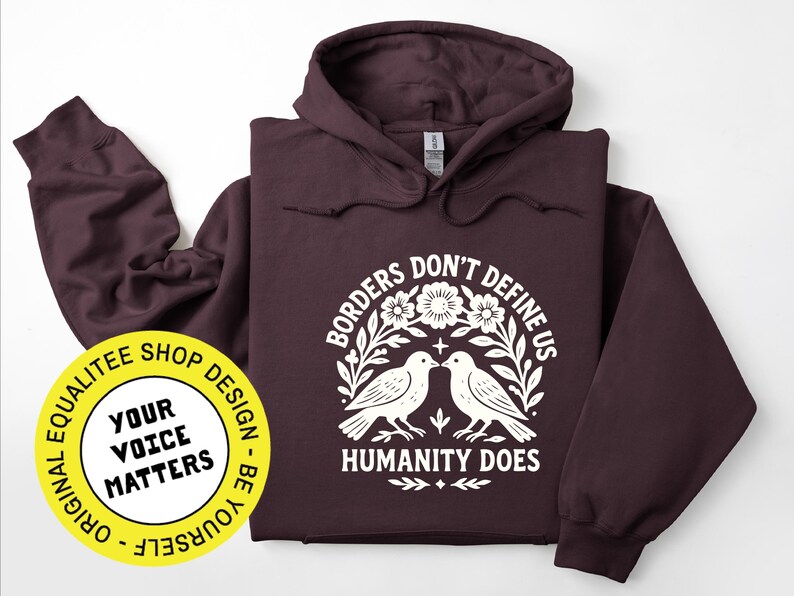 Pode incluir: Um moletom vinho com um gr&aacute;fico branco que diz "Borders Don't Define Us, Humanity Does". O design inclui dois p&aacute;ssaros, flores e folhas. &Agrave; esquerda, h&aacute; um adesivo amarelo com o texto "Your Voice Matters".