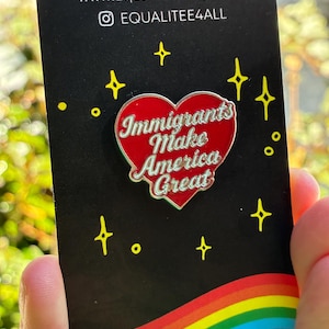 May include: A black card with a red heart-shaped pin that reads "Immigrants Make America Great." The card also features the website www.EqualiTeeShop.com and the Instagram handle @EQUALITEE4ALL. The card has a rainbow design at the bottom.