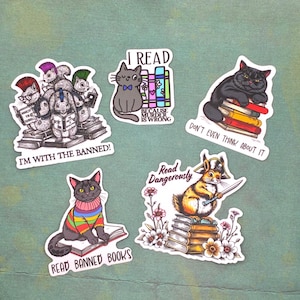 May include: A collection of five stickers featuring cats and squirrels reading books. The stickers have text that reads "I'm with the banned!", "I read because murder is wrong", "Don't even think about it", and "Read banned books".