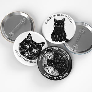 May include: A collection of black and white cat-themed button pins. One pin features a black cat with the text "Don't let the cat out or ICE in." Another says "Feeling Fractious." A third pin reads "We're all in this together like hostages."