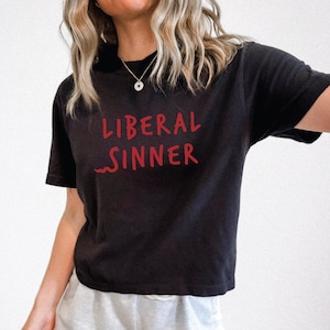 May include: Black t-shirt with the red text "LIBERAL SINNER". The shirt has short sleeves and a crew neck. The text is in a handwritten style. The model is wearing a silver necklace and light gray shorts.