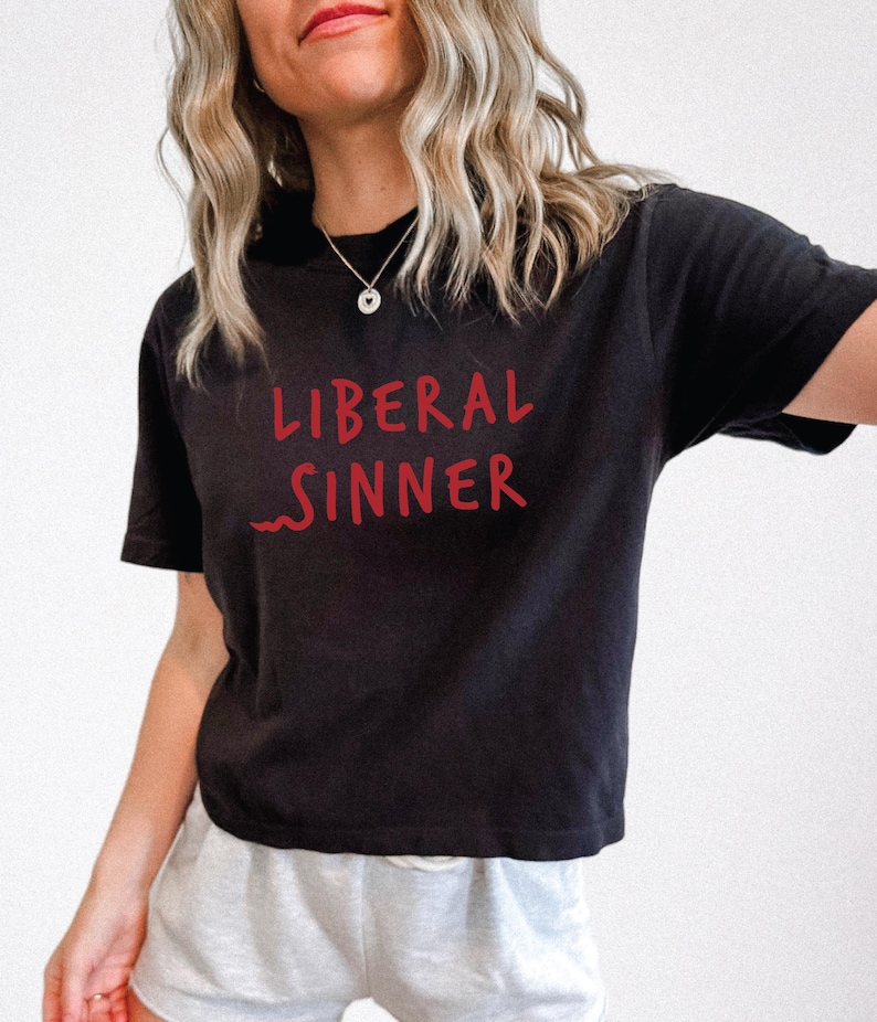 May include: Black t-shirt with the red text "LIBERAL SINNER". The shirt has short sleeves and a crew neck. The text is in a handwritten style. The model is wearing a silver necklace and light gray shorts.