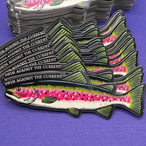 May include: A stack of fish-shaped stickers with a black outline and a colorful design. The fish have green fins, pink bellies with black spots, and the text "SWIM AGAINST THE CURRENT" in white. The background is a solid purple.
