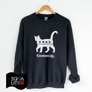 K&ouml;nnte beinhalten: Ein schwarzer Pullover mit einer wei&szlig;en Silhouette einer Katze mit f&uuml;nf Sternen auf dem R&uuml;cken. Der Text "Democat" ist unter der Katze aufgedruckt.
