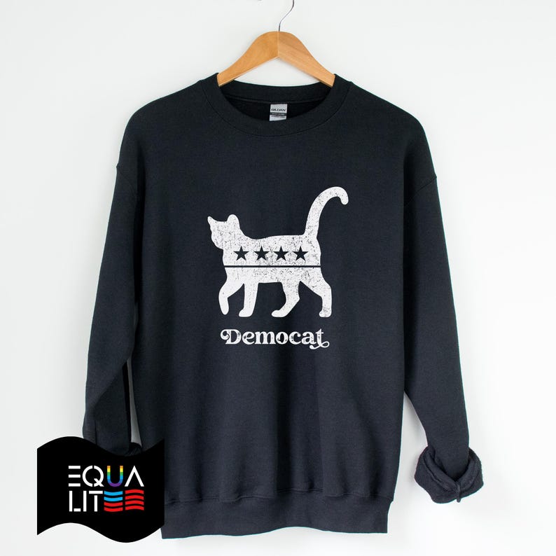 K&ouml;nnte beinhalten: Ein schwarzer Pullover mit einer wei&szlig;en Silhouette einer Katze mit f&uuml;nf Sternen auf dem R&uuml;cken. Der Text "Democat" ist unter der Katze aufgedruckt.