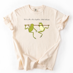 May include: Cream-colored t-shirt featuring a cartoon illustration of two green frogs hanging from a brown stick. The text above the frogs reads, "We're all in this together. Like hostages."