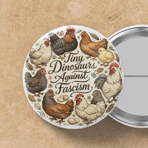 May include: A round button with a cream background featuring illustrations of chickens and the text "Tiny Dinosaurs Against Fascism" in a decorative font. The button has a metal backing.