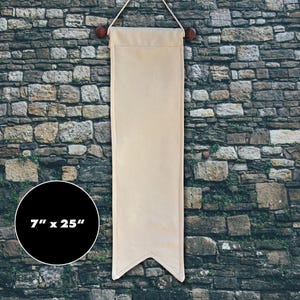 May include: A beige fabric wall banner with a pointed bottom edge, suspended from a wooden rod with two round finials and a hanging cord. The banner measures 7 inches by 25 inches as indicated by the text on the circular black graphic.