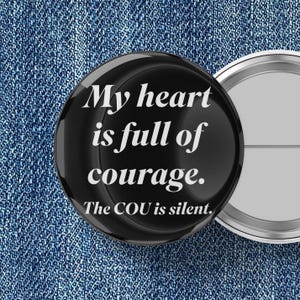 May include: A black button with the text "My heart is full of courage. The COU is silent." in white. The button has a glossy finish and a silver metal backing. The background is a blue denim fabric.