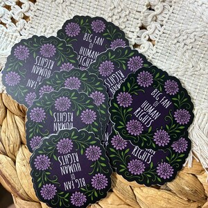 May include: A collection of circular stickers with a dark purple background, featuring a scalloped edge, green leafy vines, purple flower accents, and the white text "BIG FAN OF HUMAN RIGHTS" printed in the center of each individual sticker.