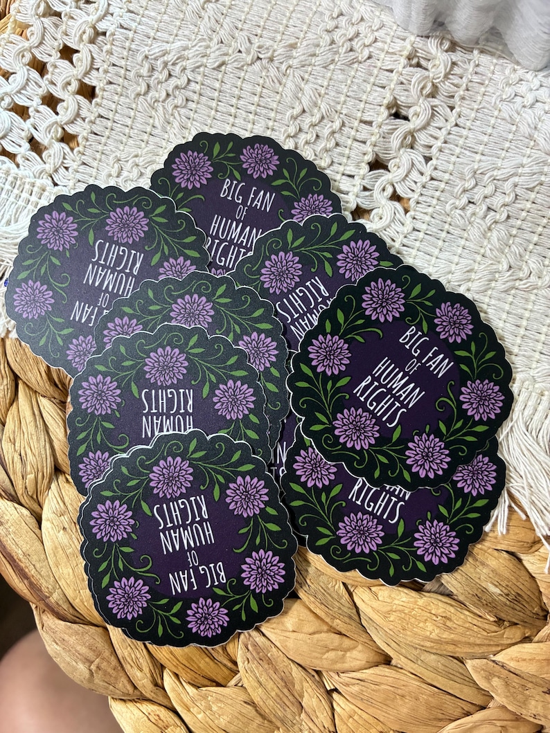 May include: A collection of circular stickers with a dark purple background, featuring a scalloped edge, green leafy vines, purple flower accents, and the white text "BIG FAN OF HUMAN RIGHTS" printed in the center of each individual sticker.