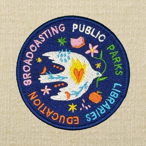 Pode incluir: Patch bordado circular azul marinho. Apresenta uma pomba branca com uma chama amarela e laranja, rodeada por motivos florais e estelares coloridos. O texto "BROADCASTING PUBLIC PARKS LIBRARIES EDUCATION" &eacute; bordado ao redor da borda.