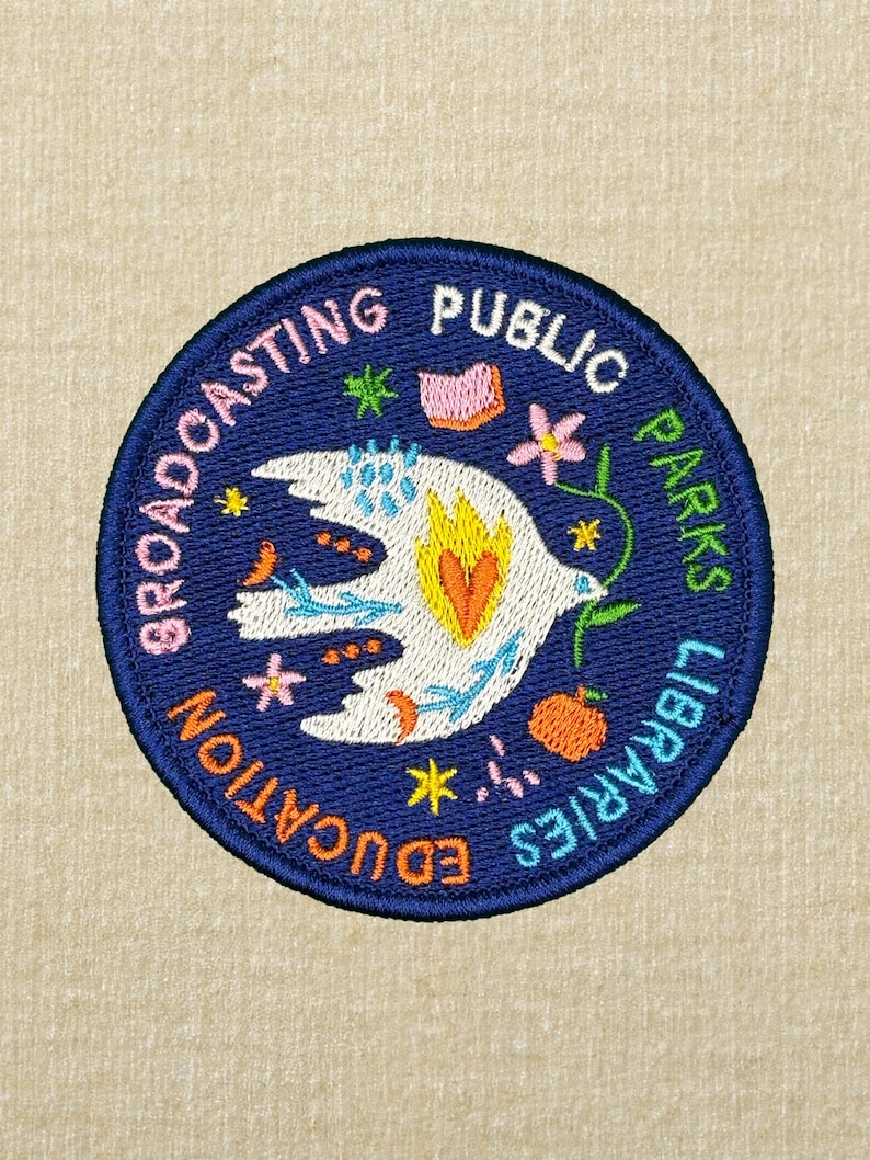 Pode incluir: Patch bordado circular azul marinho. Apresenta uma pomba branca com uma chama amarela e laranja, rodeada por motivos florais e estelares coloridos. O texto "BROADCASTING PUBLIC PARKS LIBRARIES EDUCATION" &eacute; bordado ao redor da borda.