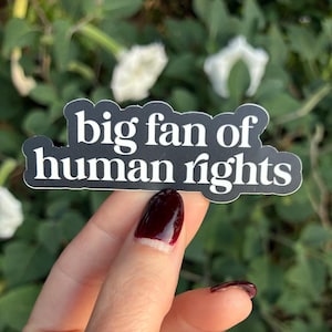 May include: A black sticker with white text that reads "big fan of human rights." The sticker is held by a hand with dark red nail polish. The background is blurred green foliage and white flowers.