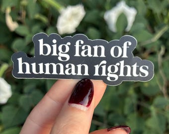 May include: A black sticker with white text that reads "big fan of human rights." The sticker is held by a hand with dark red nail polish. The background is blurred green foliage and white flowers.