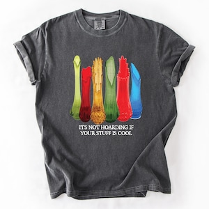 May include: Dark gray t-shirt featuring a row of colorful, vintage-style vases in green, red, yellow, and blue. The text "IT'S NOT HOARDING IF YOUR STUFF IS COOL" is printed below the vases. The shirt has rolled-up sleeves.
