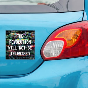May include: A square car magnet with the text "THE REVOLUTION WILL NOT BE TELEVISED" in white, set against a colorful, glitch-art background. The magnet is affixed to the rear of a light blue car, near the taillight.