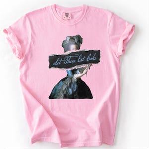 Pu&ograve; includere: T-shirt rosa chiaro con una grafica di una figura storica che tiene in mano un cartello con la scritta "Let Them Eat Cake". La figura indossa un abito blu e un grande cappello ornato. Le maniche sono arrotolate.