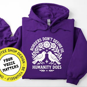 Pode incluir: Um moletom roxo com um gr&aacute;fico branco que diz "Borders Don't Define Us, Humanity Does" com duas pombas e elementos florais. Um logotipo circular amarelo com o texto "Your Voice Matters" tamb&eacute;m &eacute; vis&iacute;vel.