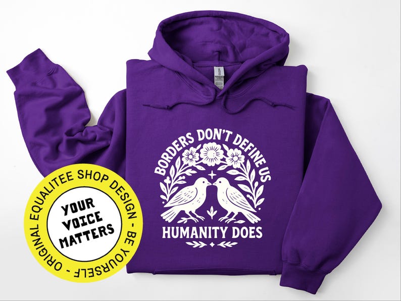 Pode incluir: Um moletom roxo com um gr&aacute;fico branco que diz "Borders Don't Define Us, Humanity Does" com duas pombas e elementos florais. Um logotipo circular amarelo com o texto "Your Voice Matters" tamb&eacute;m &eacute; vis&iacute;vel.