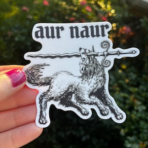 May include: A white sticker with a black illustration of a wolf holding a sword in its mouth, with the words "aur naur" above it. The sticker has a unique shape and is held against a blurred green background.