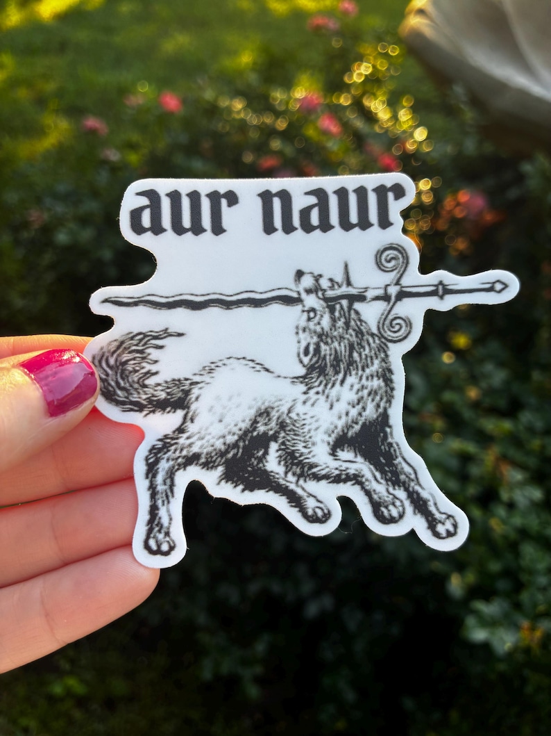 May include: A white sticker with a black illustration of a wolf holding a sword in its mouth, with the words "aur naur" above it. The sticker has a unique shape and is held against a blurred green background.