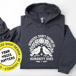 Pode incluir: Moletom cinza escuro com estampa branca: "Borders Don't Define Us, Humanity Does". O design apresenta dois p&aacute;ssaros e elementos florais. Um adesivo amarelo com "Your Voice Matters" &eacute; vis&iacute;vel. O moletom tem capuz.