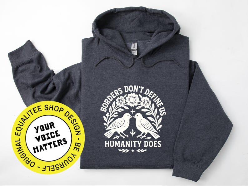 Pode incluir: Moletom cinza escuro com estampa branca: "Borders Don't Define Us, Humanity Does". O design apresenta dois p&aacute;ssaros e elementos florais. Um adesivo amarelo com "Your Voice Matters" &eacute; vis&iacute;vel. O moletom tem capuz.