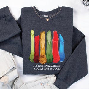 May include: A dark gray sweatshirt featuring a colorful graphic of stylized vases in shades of green, red, orange, and blue. The text "IT'S NOT HOARDING IF YOUR STUFF IS COOL" is printed below the vases. A gold necklace is visible.