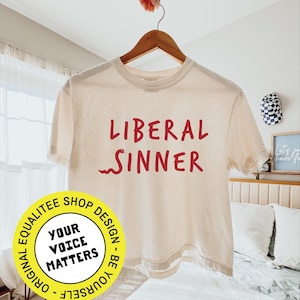 May include: Cream-colored t-shirt with the words "LIBERAL SINNER" in red script. The shirt is hanging on a wooden hanger. A round yellow sticker with the text "YOUR VOICE MATTERS" is in the lower left corner.