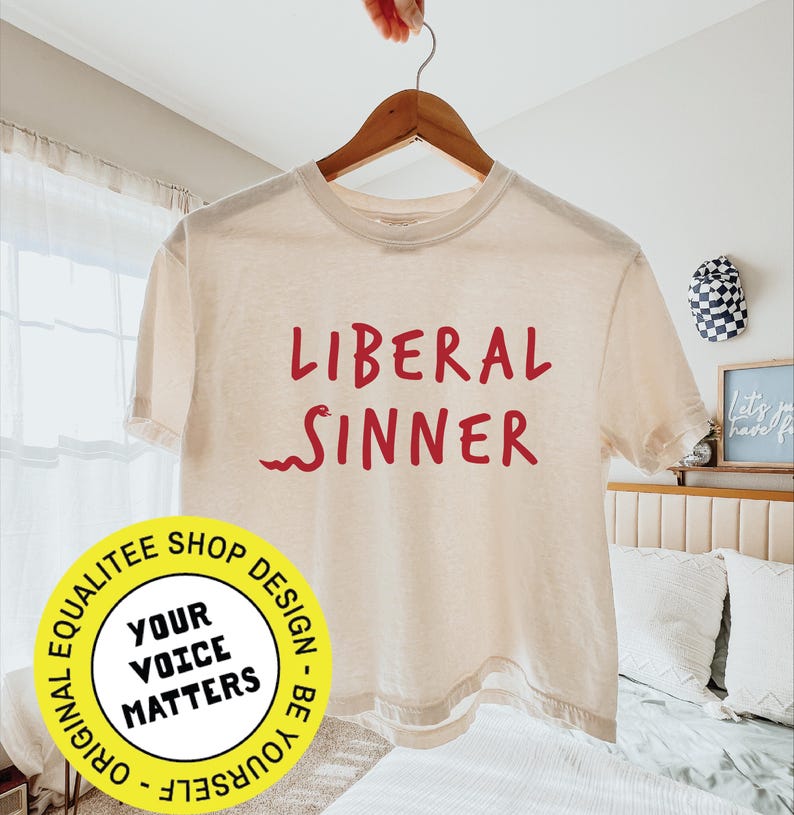 May include: Cream-colored t-shirt with the words "LIBERAL SINNER" in red script. The shirt is hanging on a wooden hanger. A round yellow sticker with the text "YOUR VOICE MATTERS" is in the lower left corner.
