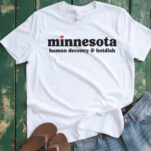 May include: White t-shirt with black text that reads "minnesota human decency & hotdish" with a small red heart above the word "minnesota".