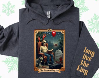 May include: A dark gray hooded sweatshirt with a graphic print of a skeleton sitting in a chair reading a book. The graphic is in a vintage style with a red balloon and the text "The Stephen King Fan" at the bottom. The right sleeve has the text "Long Live The King" printed in a gold color.