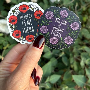 May include: Two round stickers with floral designs and text. One sticker has red poppies and the text "TU LUCHA ES MI LUCHA". The other has pink flowers and the text "BIG FAN OF HUMAN RIGHTS".