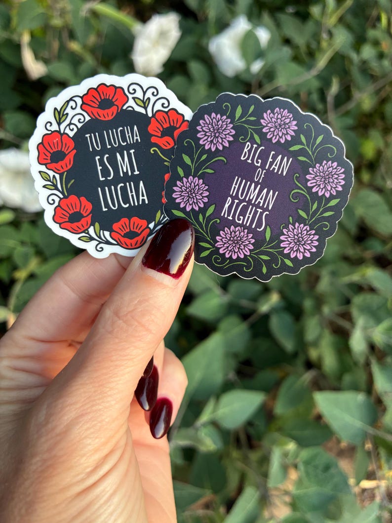 May include: Two round stickers with floral designs and text. One sticker has red poppies and the text "TU LUCHA ES MI LUCHA". The other has pink flowers and the text "BIG FAN OF HUMAN RIGHTS".