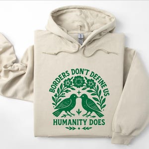Pode incluir: Um moletom bege com um gr&aacute;fico verde que diz "Borders Don't Define Us, Humanity Does". O gr&aacute;fico apresenta dois p&aacute;ssaros voltados um para o outro, cercados por flores e folhas. O moletom tem capuz com cord&atilde;o e mangas compridas.
