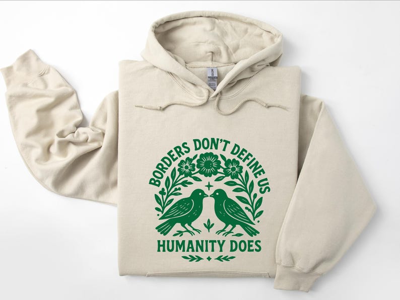 Pode incluir: Um moletom bege com um gr&aacute;fico verde que diz "Borders Don't Define Us, Humanity Does". O gr&aacute;fico apresenta dois p&aacute;ssaros voltados um para o outro, cercados por flores e folhas. O moletom tem capuz com cord&atilde;o e mangas compridas.