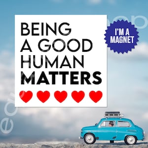 May include: White square magnet with black text that reads "Being a good human matters" with five red hearts below the text. The magnet has a blue starburst sticker with the text "I'm a magnet" in white.