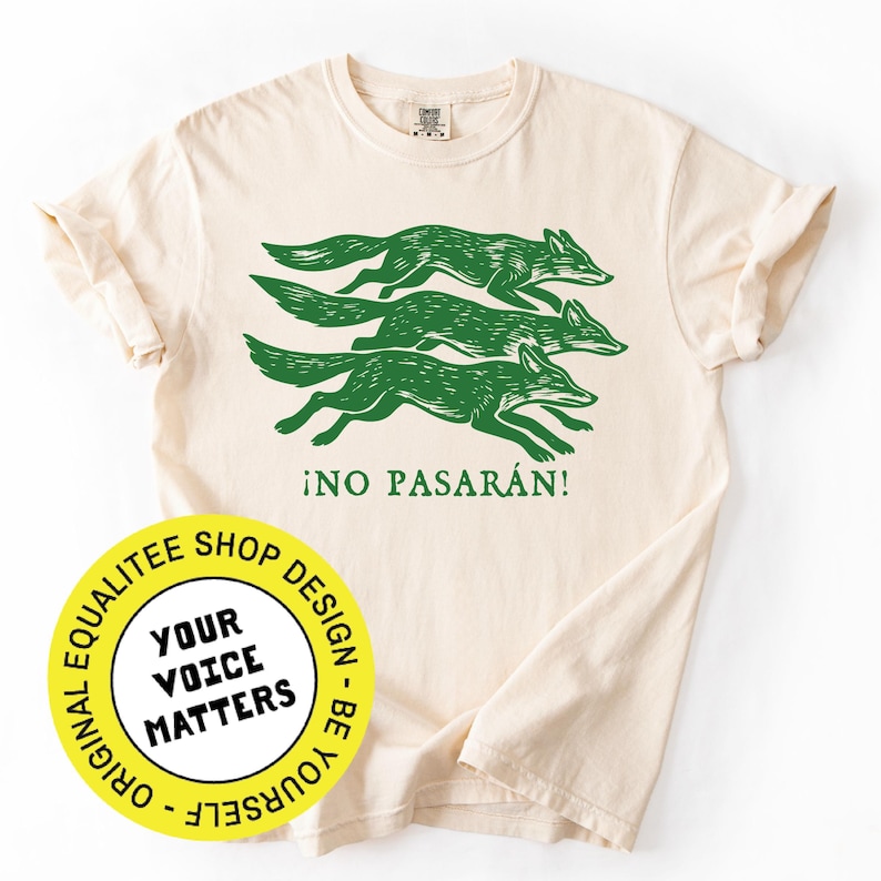 May include: Cream-colored t-shirt featuring a green graphic of three running foxes and the Spanish phrase "&iexcl;NO PASAR&Aacute;N!". A yellow circular logo with the text "YOUR VOICE MATTERS" is also visible.