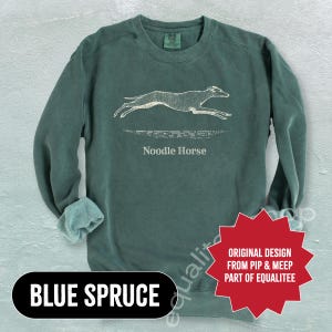 Pode incluir: Camisola azul abeto com um gr&aacute;fico branco de um galgo a correr e as palavras "Noodle Horse". A camisola tamb&eacute;m tem as palavras "Blue Spruce" num oval preto e uma estrela vermelha com texto.