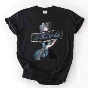 Pu&ograve; includere: T-shirt nero con ritratto storico e la scritta "Let Them Eat Cake". Il ritratto raffigura una donna con un abito blu e una grande parrucca bianca. La maglietta &egrave; realizzata in materiale morbido, adatta all'uso quotidiano.