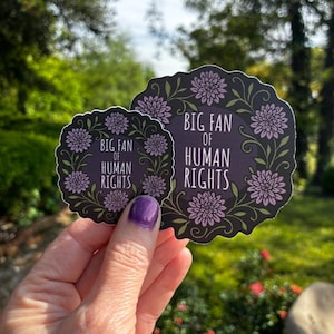 May include: Two stickers with a floral design and the text "BIG FAN OF HUMAN RIGHTS." The stickers are dark purple with a black border and feature light purple flowers and green leaves. The stickers are held up in front of a blurred green background.