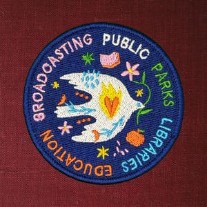 Pode incluir: Um patch redondo bordado com fundo azul marinho e texto branco que diz "Broadcasting Public Parks Libraries Education". O patch apresenta uma pomba branca com um cora&ccedil;&atilde;o amarelo e laranja, rodeada por desenhos florais e de estrelas coloridas.