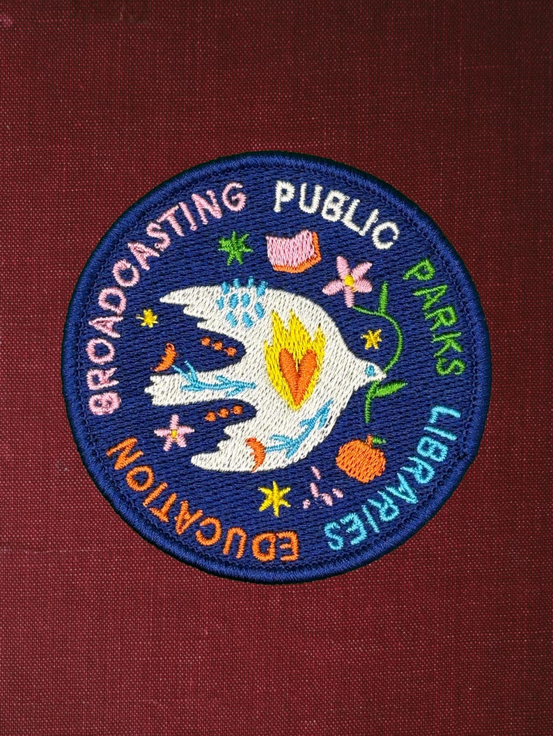 Pode incluir: Um patch redondo bordado com fundo azul marinho e texto branco que diz "Broadcasting Public Parks Libraries Education". O patch apresenta uma pomba branca com um cora&ccedil;&atilde;o amarelo e laranja, rodeada por desenhos florais e de estrelas coloridas.